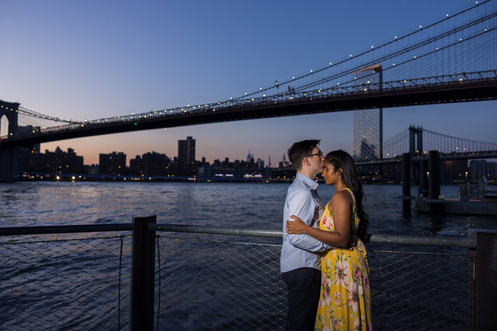 Blue hour Brooklyn Bridge engagement photos in DUMBO, with Sreetha and Demitri embracing along the waterfront as the Manhattan Bridge lights reflect across the East River and NYC skyline.