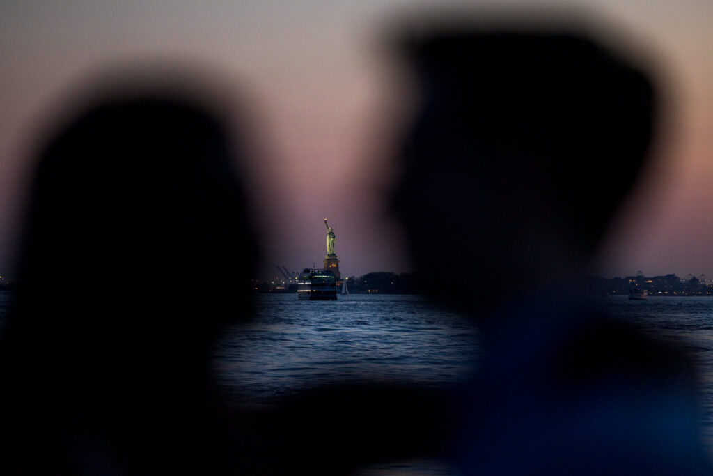 Romantic NYC engagement photo at twilight with the Statue of Liberty illuminated across the water, framed by a soft silhouette of the couple during their waterfront session.
