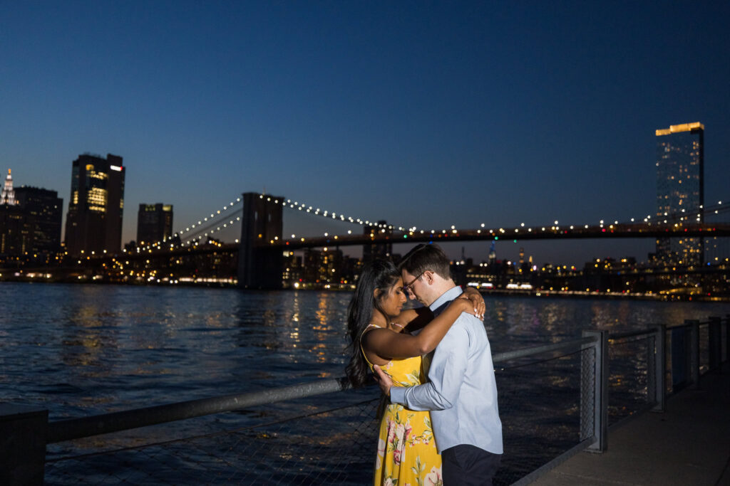 Nighttime Brooklyn Bridge Park engagement photo with Sreetha and Demitri embracing along the waterfront, Brooklyn Bridge illuminated behind them and city lights reflecting across the East River.