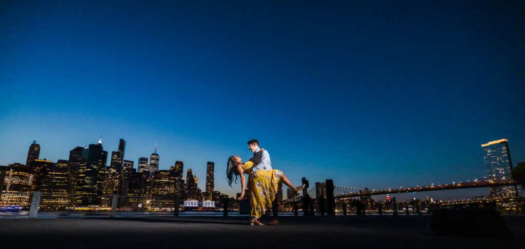 Couple in a dramatic dip pose along the Brooklyn waterfront at night, with the Manhattan skyline and Brooklyn Bridge illuminated behind them for stunning NYC engagement photos after dark.