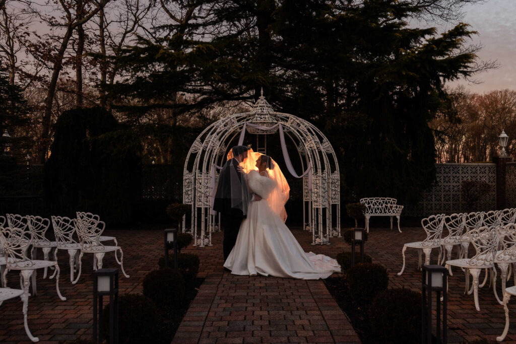 Bride and groom sharing a romantic sunset kiss beneath the garden arbor at an Estate at East Wind wedding, captured during a winter evening on Long Island.