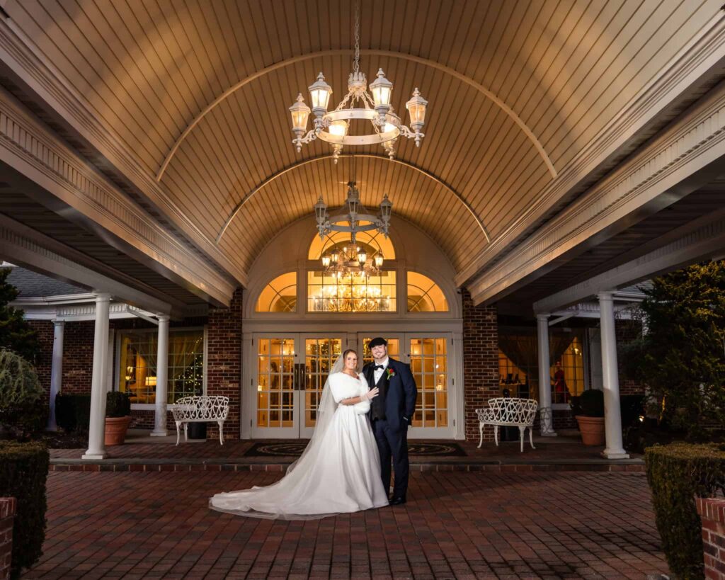 Bride and groom posing beneath the grand covered entrance of the Estate at East Wind wedding venue, highlighting the illuminated exterior and refined Long Island estate architecture.