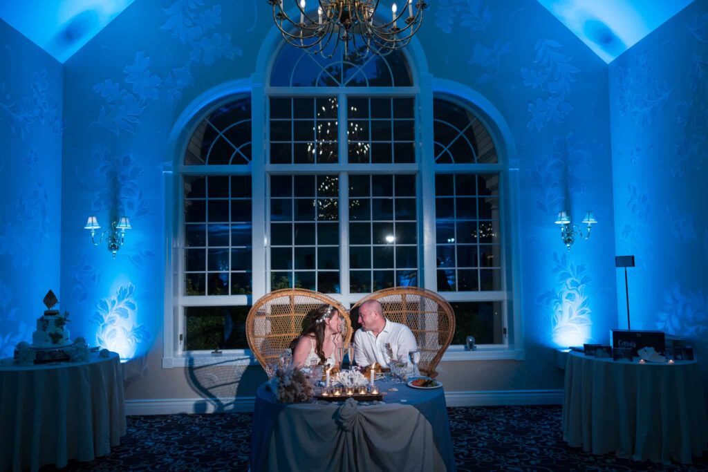 Bride and groom seated at a sweetheart table during an Estate at East Wind wedding, framed by arched windows and blue uplighting inside the elegant Long Island reception space.
