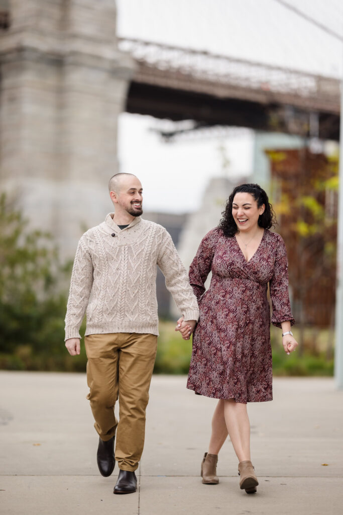 Couple walking hand in hand beneath the Manhattan Bridge during fall engagement photos in DUMBO, captured during an early morning engagement session in NYC with soft November tones.