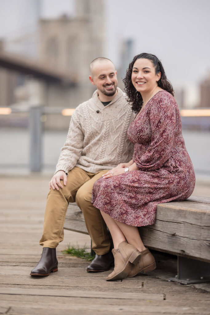 Couple seated along the Brooklyn waterfront during fall engagement photos in DUMBO, with the Brooklyn Bridge softly blurred behind them in gentle November morning light.