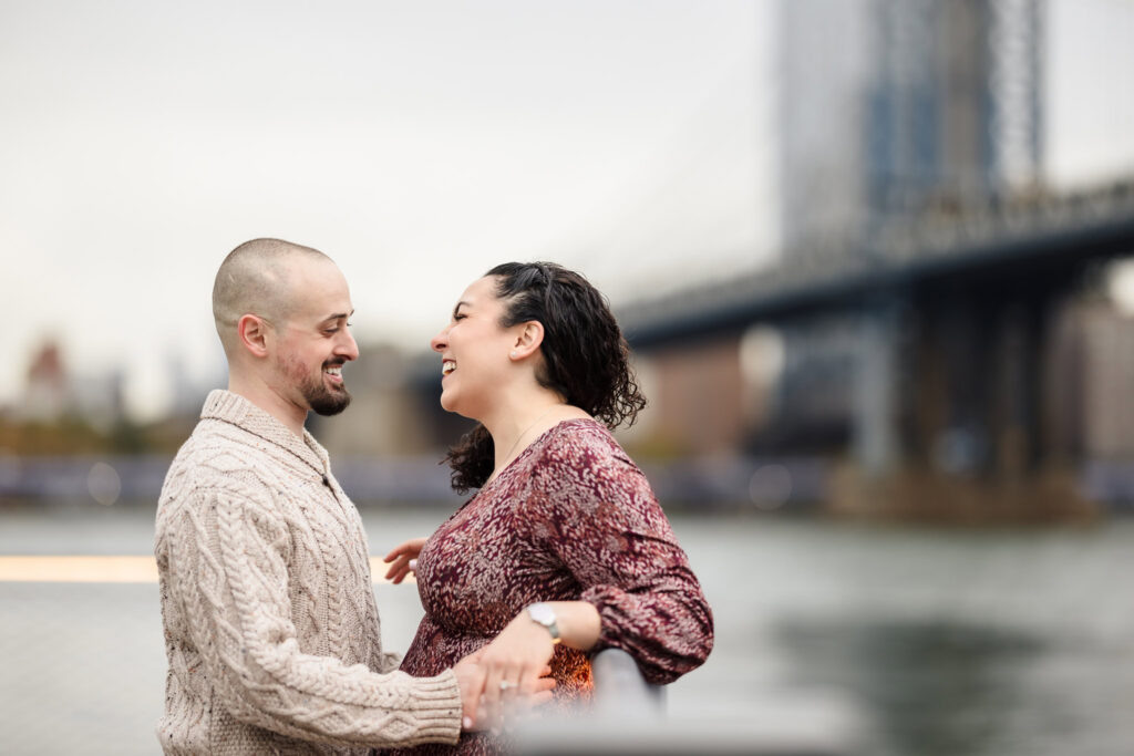 Couple laughing together along the Brooklyn waterfront during fall engagement photos in DUMBO, with the Manhattan Bridge softly blurred behind them in November morning light.