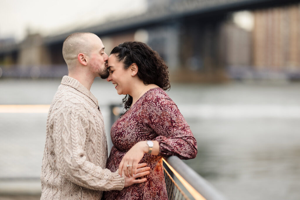 Groom kissing bride’s forehead during fall engagement photos in DUMBO, captured along the Brooklyn waterfront with the Manhattan Bridge in soft November morning light.