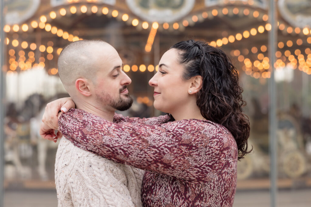 Couple embracing in front of a vintage carousel during fall engagement photos in DUMBO, with warm bokeh lights adding depth to this November engagement session in Brooklyn.