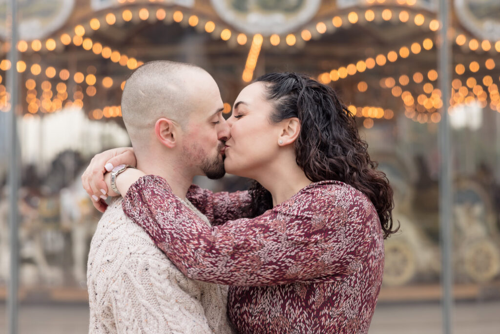 Couple sharing a kiss in front of Jane’s Carousel, with glowing lights creating a romantic November engagement session backdrop in Brooklyn.