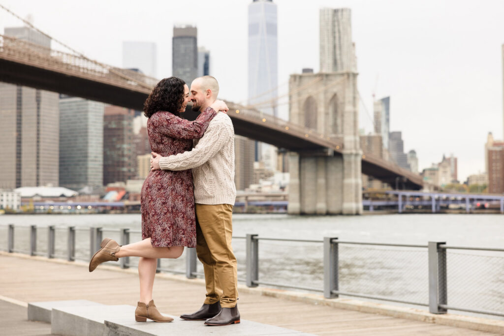 Couple embracing along the Brooklyn waterfront with the Brooklyn Bridge and Manhattan skyline behind them during fall engagement photos in DUMBO.