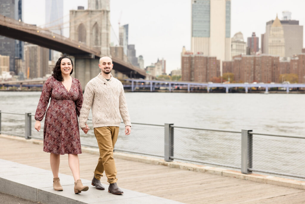 Couple walking along Brooklyn Bridge Park with the Brooklyn Bridge and Manhattan skyline behind them during a DUMBO engagement session, capturing classic Brooklyn waterfront engagement photos in DUMBO Brooklyn.
