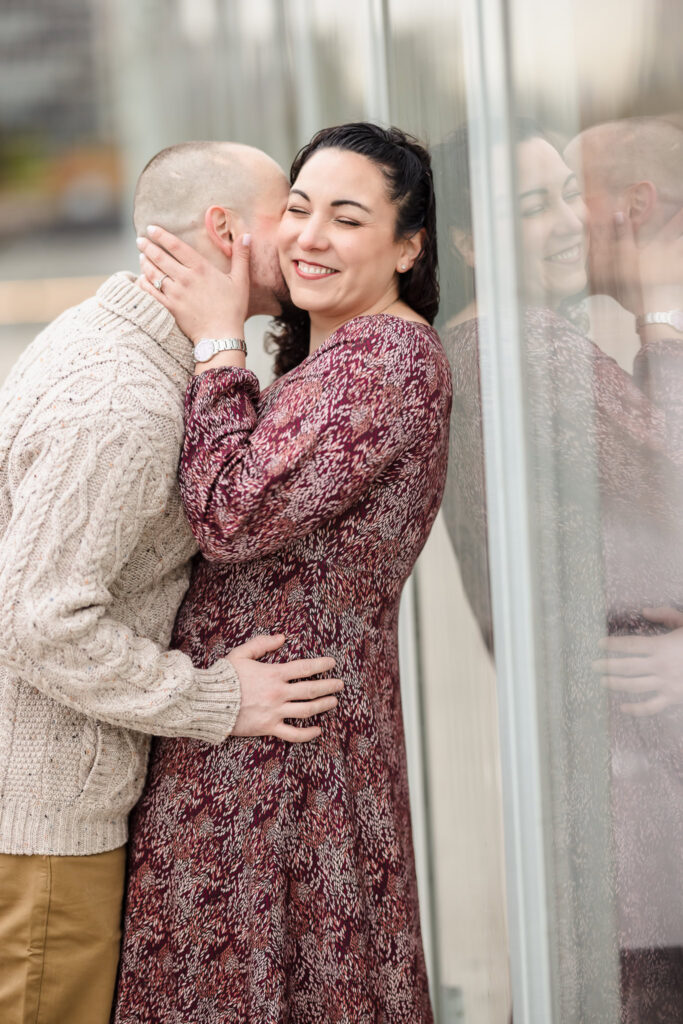 Bride smiling as her fiancé kisses her cheek, reflected in a glass facade along the Brooklyn waterfront in November.