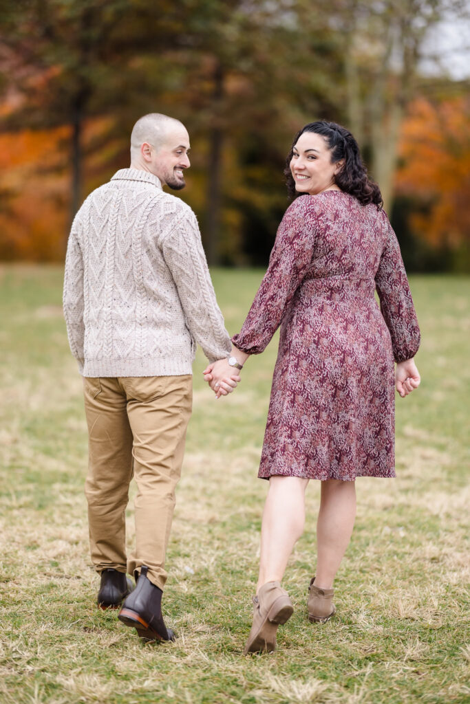 Couple holding hands and walking through autumn foliage during a November engagement session in Brooklyn, featuring golden fall tones and natural movement.