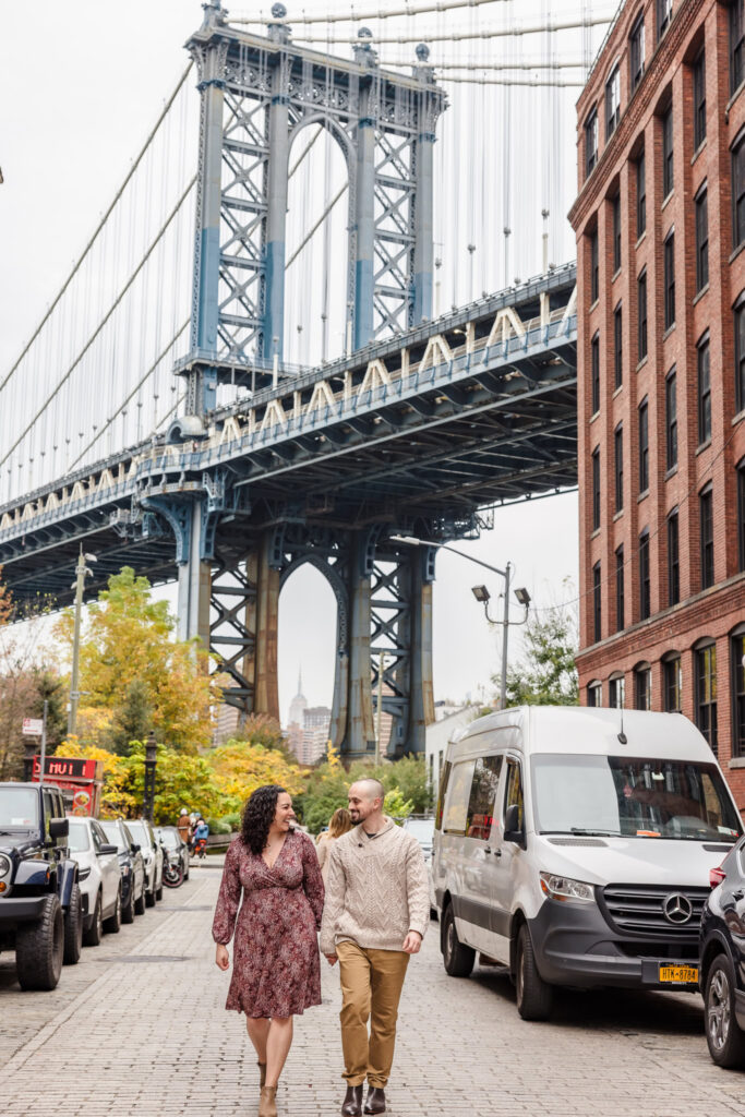Couple walking along Washington Street in DUMBO beneath the Manhattan Bridge during fall, capturing seasonal charm near iconic NYC engagement photo spots.