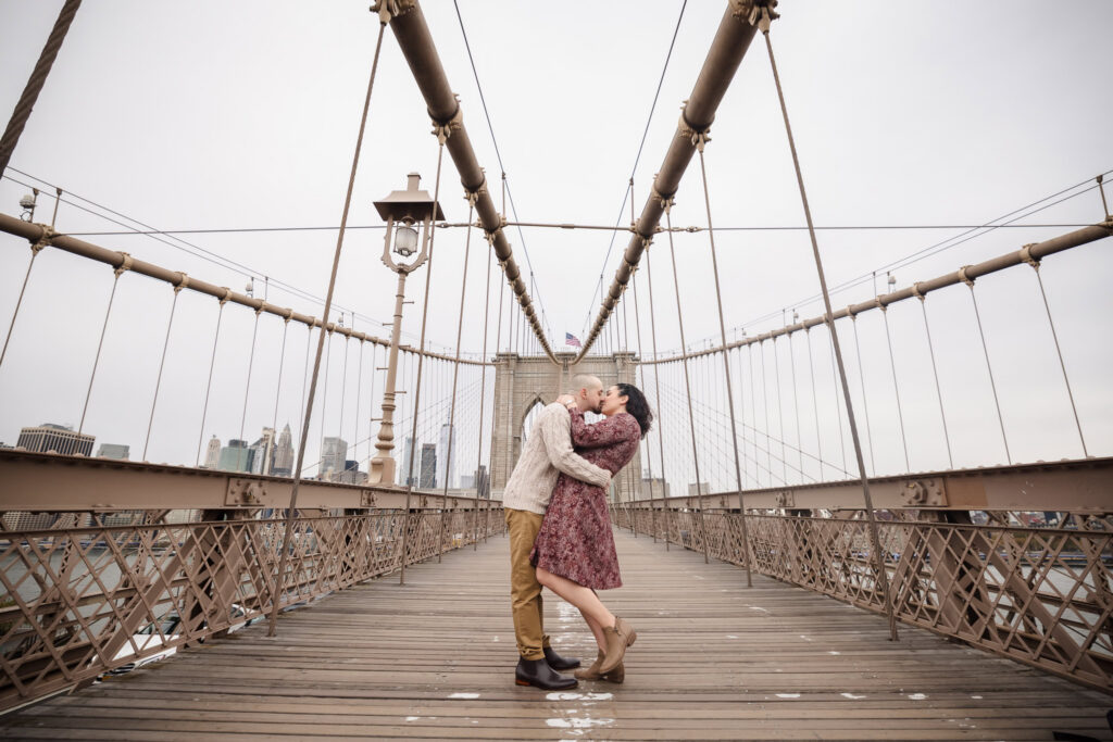 Couple kissing at the center of the Brooklyn Bridge walkway with Manhattan skyline in the background, showcasing architectural engagement photos NYC couples love near DUMBO.