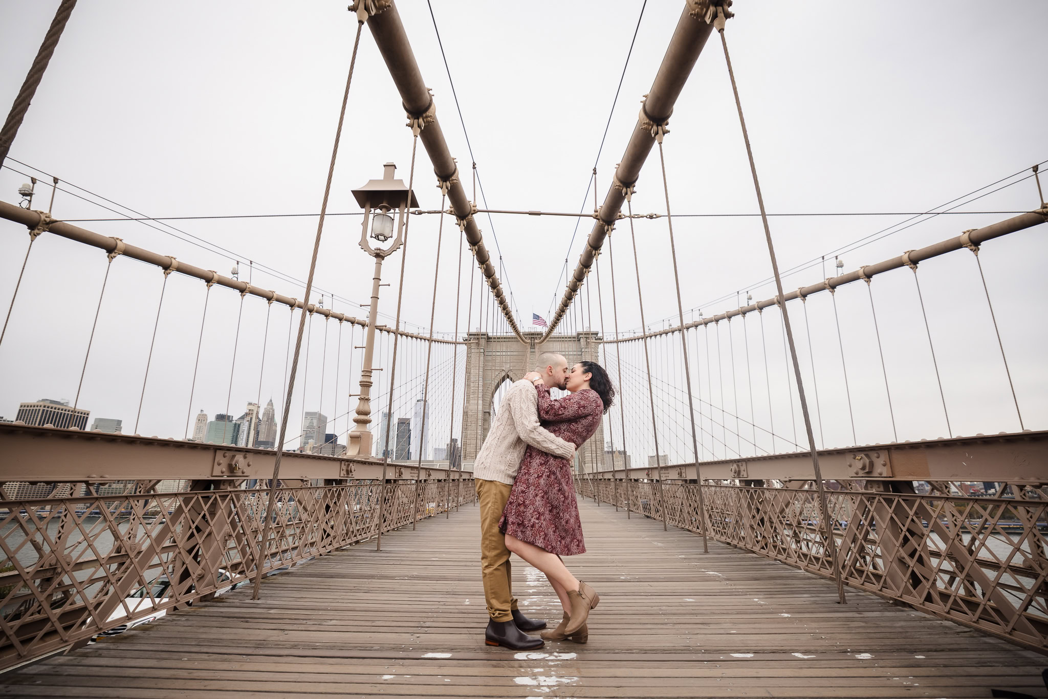 Couple kissing at the center of the Brooklyn Bridge during fall engagement photos in DUMBO, framed symmetrically with the Manhattan skyline in soft November light.