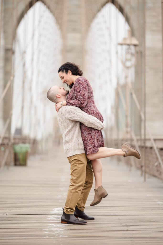 Groom lifting his bride on the Brooklyn Bridge during fall engagement photos in DUMBO, framed by the iconic stone arches in soft November morning light.