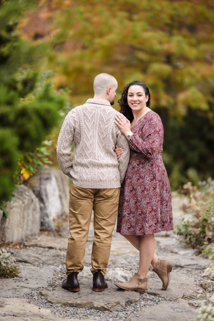 Couple standing along a garden path surrounded by golden autumn foliage during a November engagement session in Brooklyn, capturing soft fall tones and natural connection.