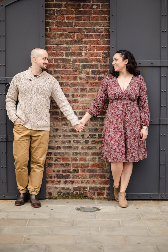 Couple holding hands against classic DUMBO brick and industrial metal doors during fall engagement photos in DUMBO, highlighting architectural textures and refined NYC portrait style.