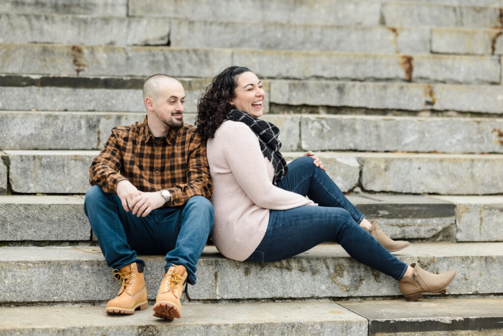 Playful couple sitting on stone steps in Brooklyn Bridge Park during fall, showing relaxed engagement session style in DUMBO.