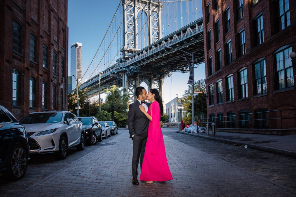 Elegant couple kissing on Washington Street in DUMBO beneath the Manhattan Bridge, capturing iconic Washington Street DUMBO photos with dramatic city architecture and cobblestone streets.