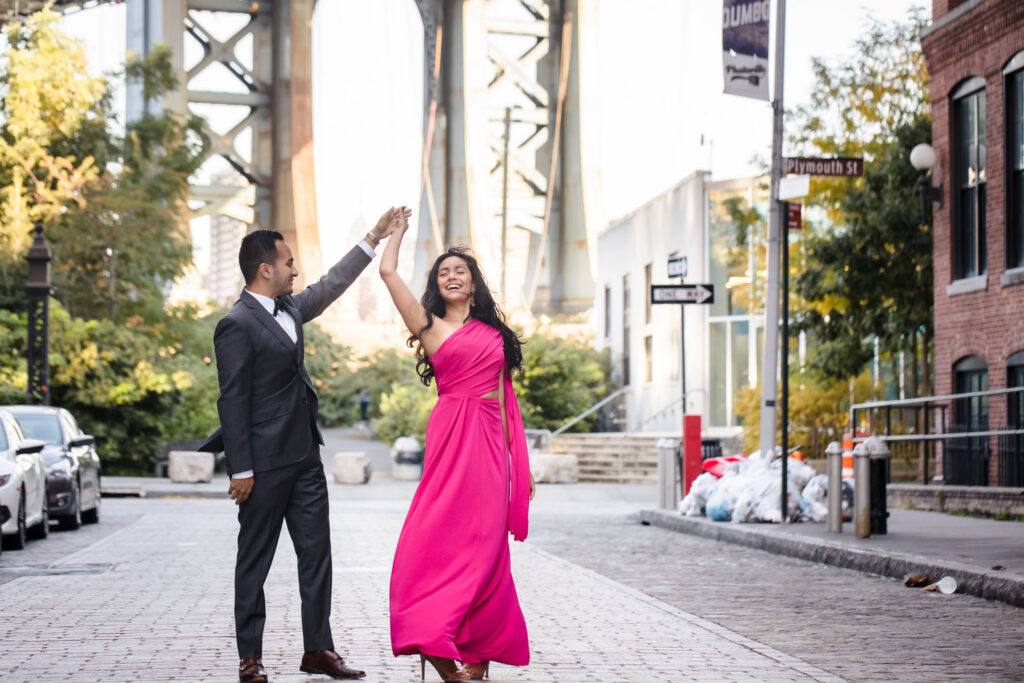 Couple dancing on Washington Street in DUMBO with the Manhattan Bridge overhead, capturing joyful Manhattan Bridge engagement photos along iconic cobblestone streets in Brooklyn.