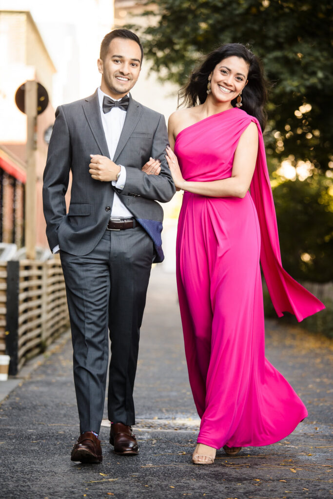 Couple walking arm in arm in DUMBO wearing a tailored gray suit and flowing fuchsia gown, demonstrating what to wear for engagement photos in DUMBO with structured silhouettes and movement in morning light Brooklyn.