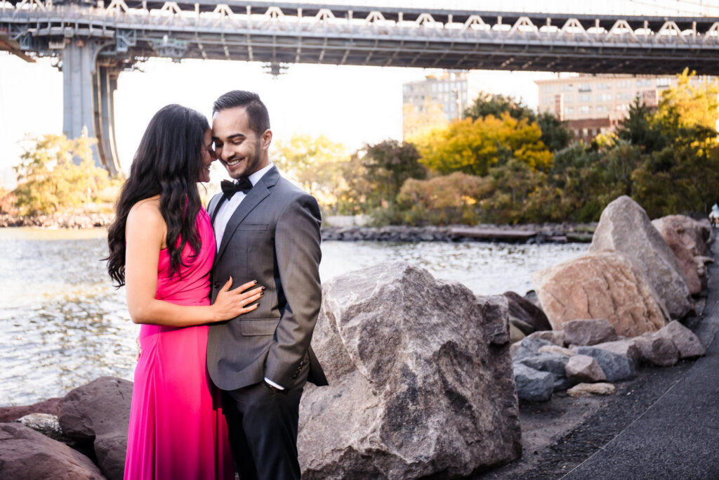 Brooklyn waterfront engagement session featuring Mahati and Winston in formal attire beneath the Manhattan Bridge, capturing luxury engagement photos in DUMBO with skyline and river views.
