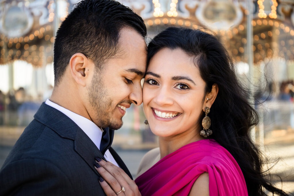 Close-up portrait at Jane’s Carousel in DUMBO showcasing formal engagement attire NYC, with a tailored suit and flowing fuchsia gown styled for luxury engagement photos Brooklyn style against a softly lit architectural backdrop.