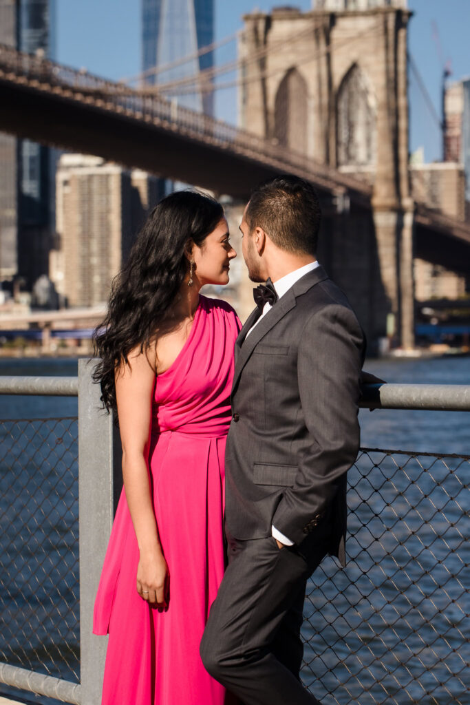 Mahati and Winston standing along the Brooklyn waterfront in DUMBO, framed by the Brooklyn Bridge and Lower Manhattan skyline, capturing refined NYC engagement portraits in formal attire.