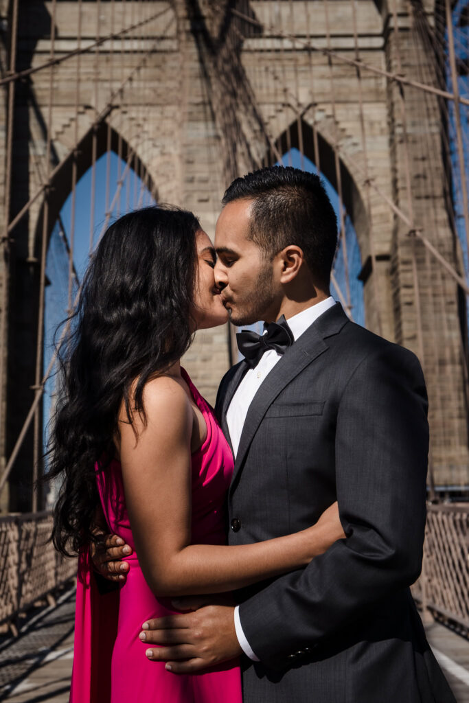 Mahati and Winston sharing a kiss on the Brooklyn Bridge during their luxury engagement session in DUMBO, framed by the bridge’s iconic stone arches and architectural lines.