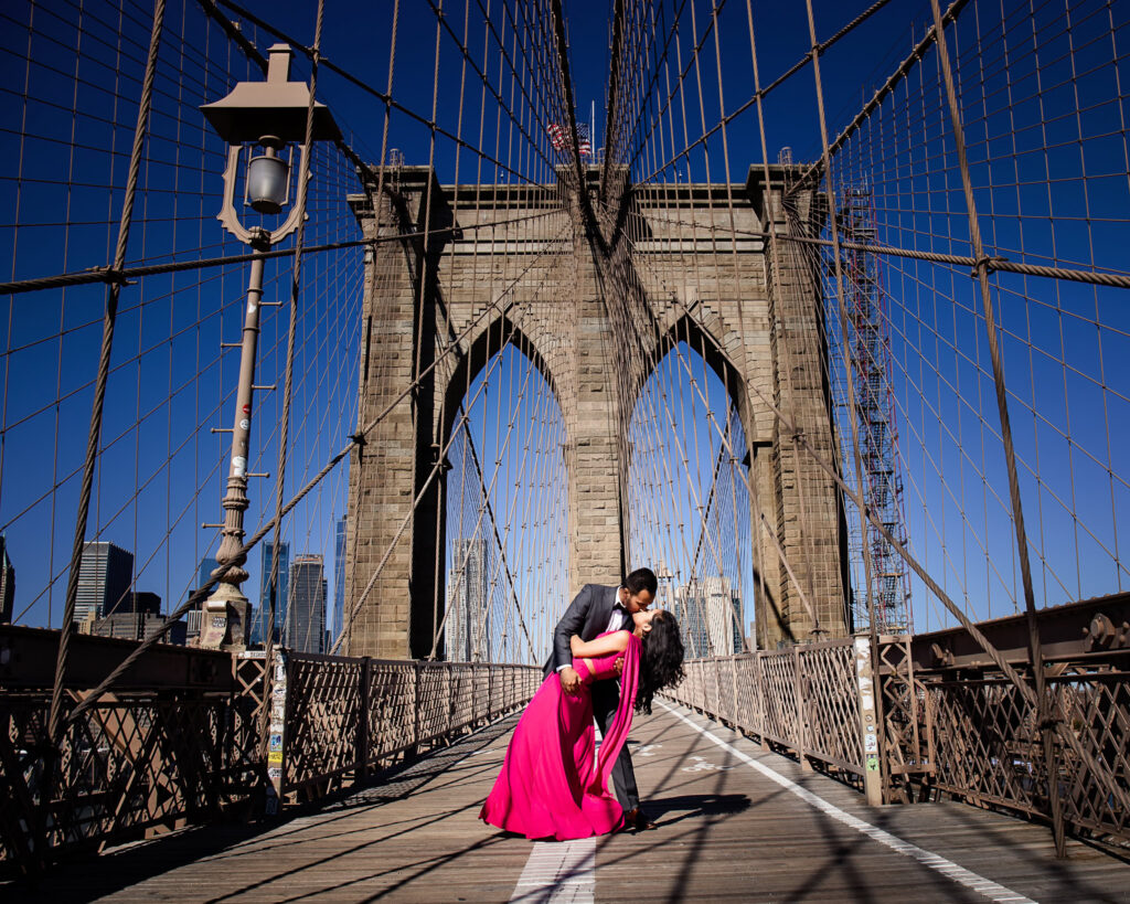 Luxury engagement photos in DUMBO capturing Mahati and Winston in a dramatic dip and kiss on the Brooklyn Bridge, framed by symmetrical architectural lines and a clear New York skyline.