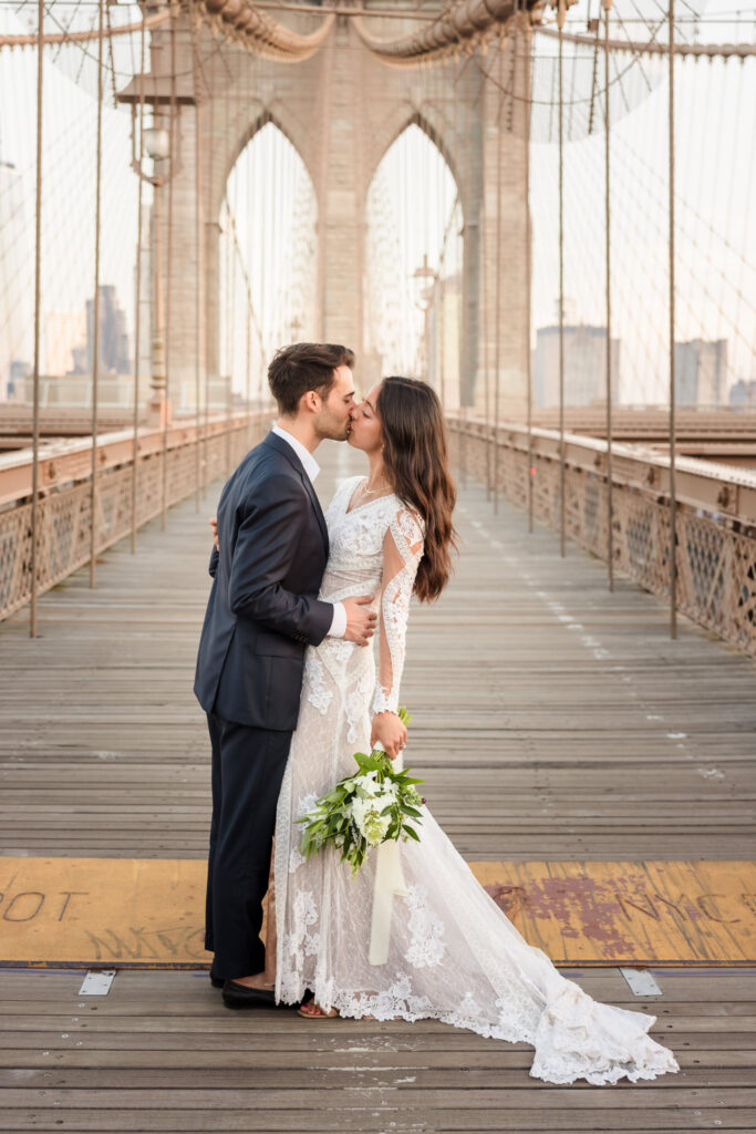 Couple kissing at sunrise on the Brooklyn Bridge during their DUMBO engagement session, framed by symmetrical bridge architecture for timeless NYC engagement photos.