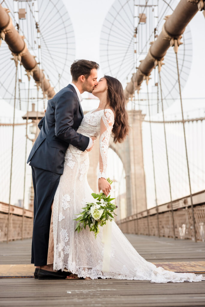 Sunrise Brooklyn Bridge wedding portrait with bride and groom kissing beneath the suspension cables, framed symmetrically by the iconic stone tower and soft July morning light.