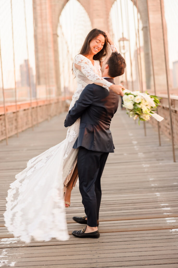 Joyful sunrise Brooklyn Bridge wedding photo of groom lifting his bride in flowing lace gown, her bouquet extended as soft July morning light illuminates the empty bridge walkway and stone arches.