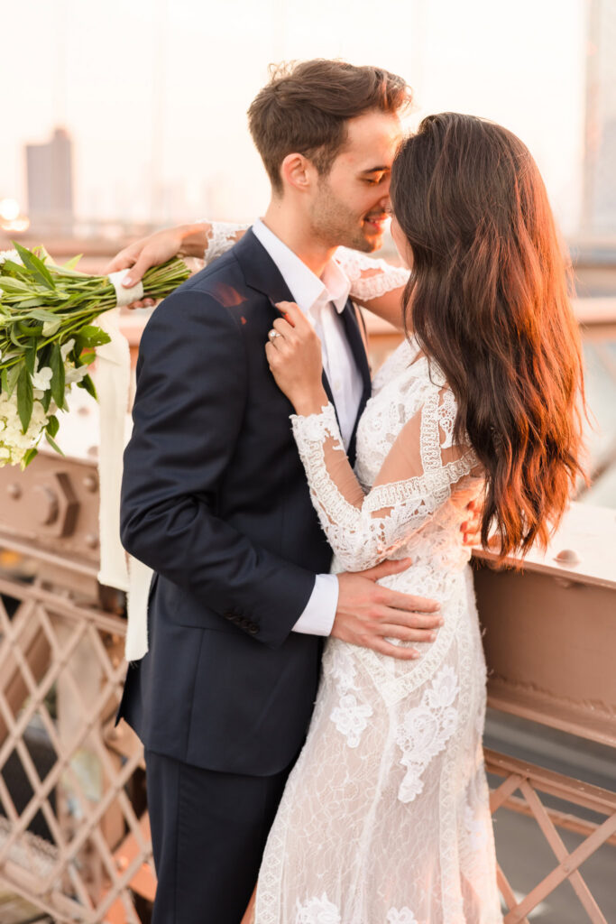 Intimate sunrise wedding portrait of bride and groom embracing along the railing, warm early morning light illuminating her lace gown and bouquet with the Manhattan skyline softly blurred behind them.