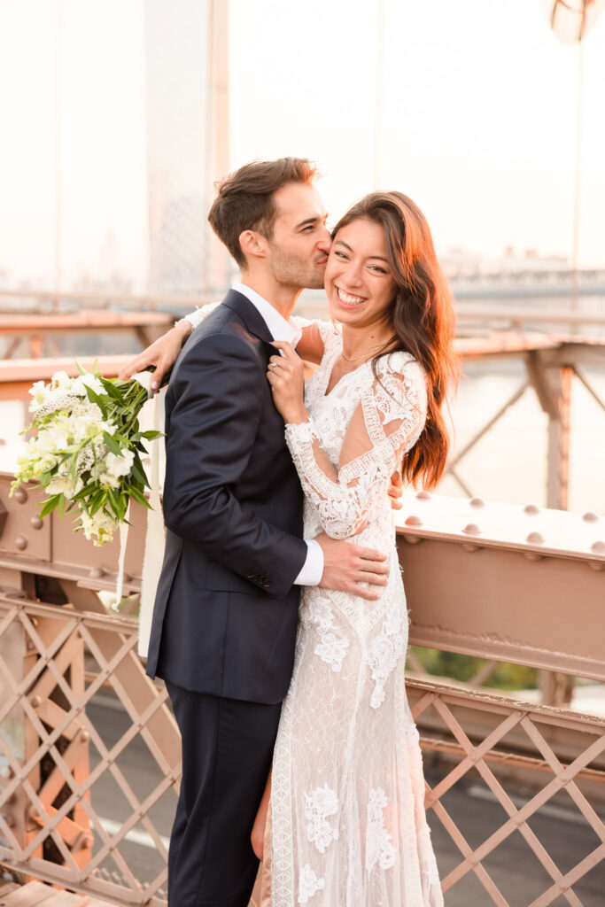 Sunrise Brooklyn Bridge wedding portrait of groom kissing bride’s cheek as she smiles toward camera, warm July morning light highlighting her lace gown and bouquet against the bridge railing and skyline.
