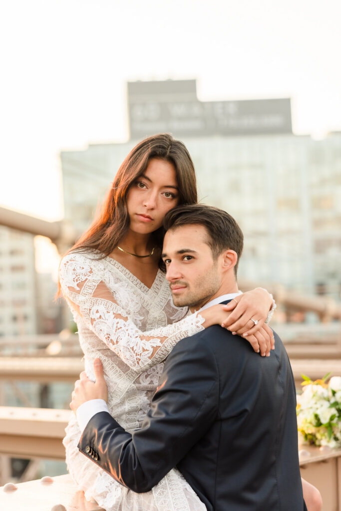 Sunrise Brooklyn Bridge wedding portrait of bride in long-sleeve lace gown seated in groom’s arms, both looking toward the camera with soft July morning light and blurred Manhattan buildings behind them.