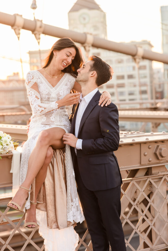 Romantic sunrise Brooklyn Bridge wedding portrait of bride sitting on the bridge railing in a lace gown, laughing with groom in navy suit as golden morning light illuminates the Manhattan skyline behind them.