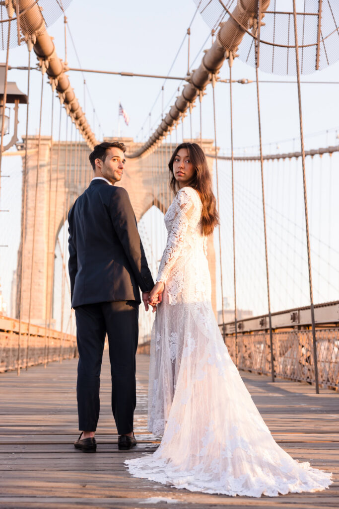 Sunrise Brooklyn Bridge wedding portrait of bride and groom holding hands and looking back toward the camera, framed by the bridge’s stone tower and suspension cables in warm early morning light.