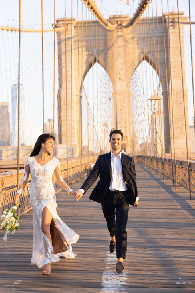 Sunrise Brooklyn Bridge wedding photo of bride and groom running hand in hand down the empty walkway, framed by the iconic stone arches and suspension cables in warm July morning light.