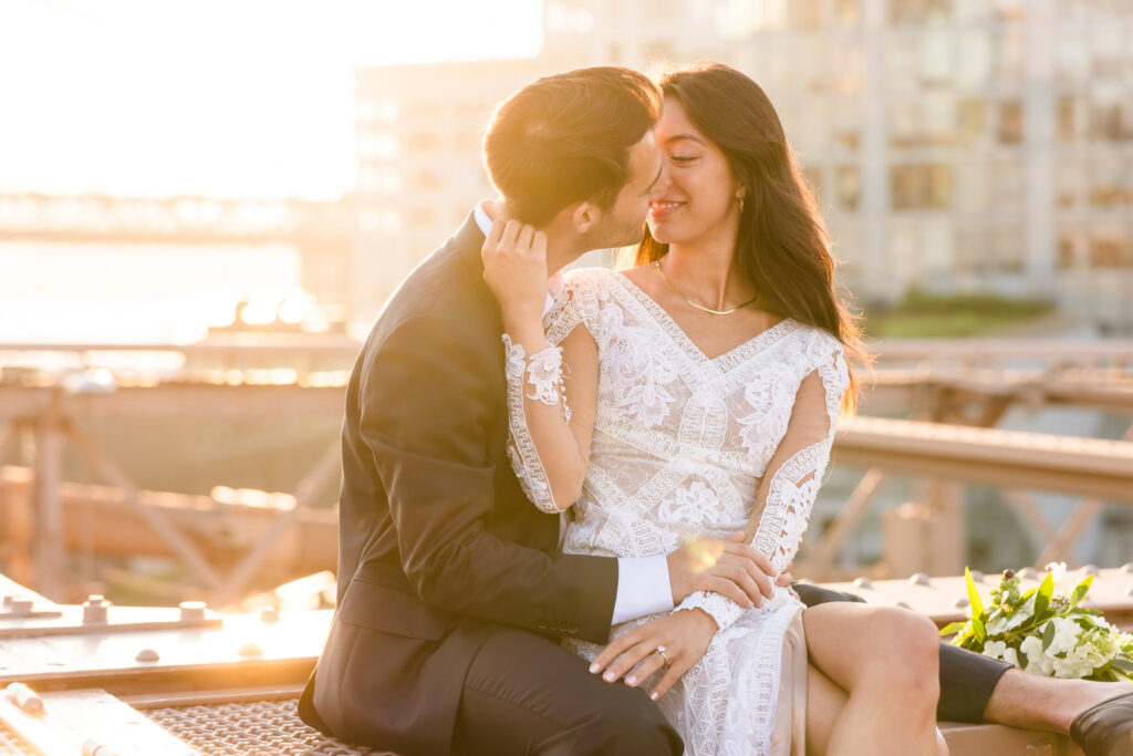 Romantic couple sitting on the Brooklyn Bridge at golden hour, sharing an intimate moment with warm sunlight and the Manhattan skyline softly glowing in the background for dreamy NYC engagement photos.