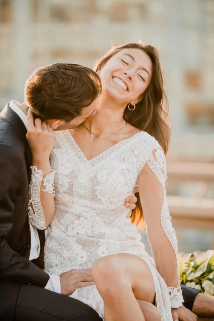Close-up wedding portrait of groom kissing bride’s neck as she smiles with eyes closed, warm July morning light illuminating her lace gown and soft Manhattan skyline background.