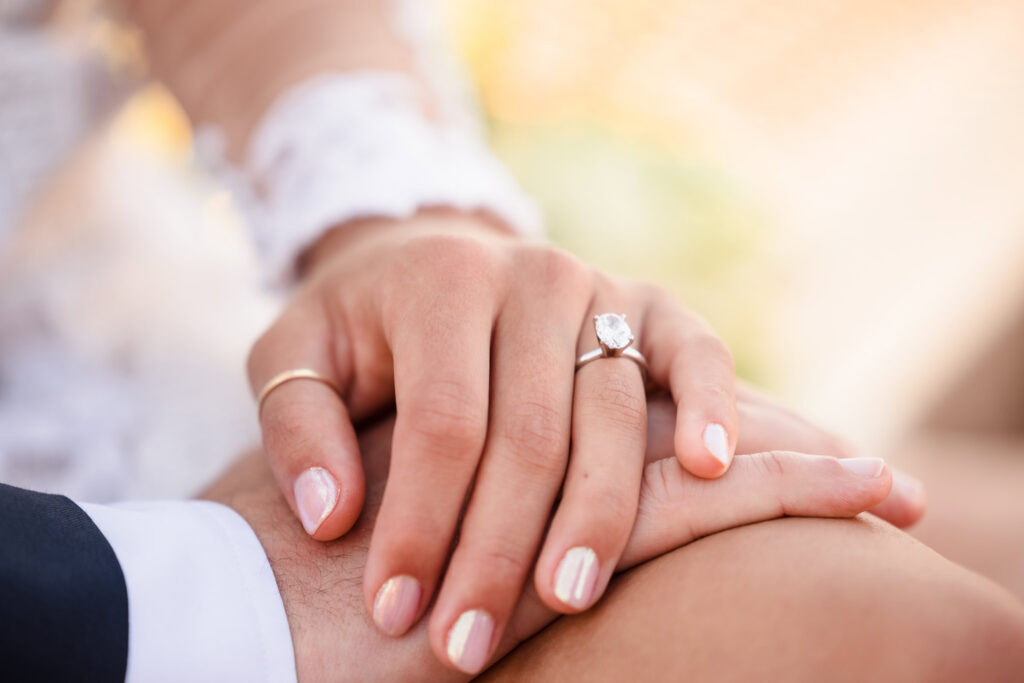 Close-up sunrise Brooklyn Bridge wedding detail of bride’s hand resting on groom’s, showcasing a solitaire diamond engagement ring in soft golden July morning light.