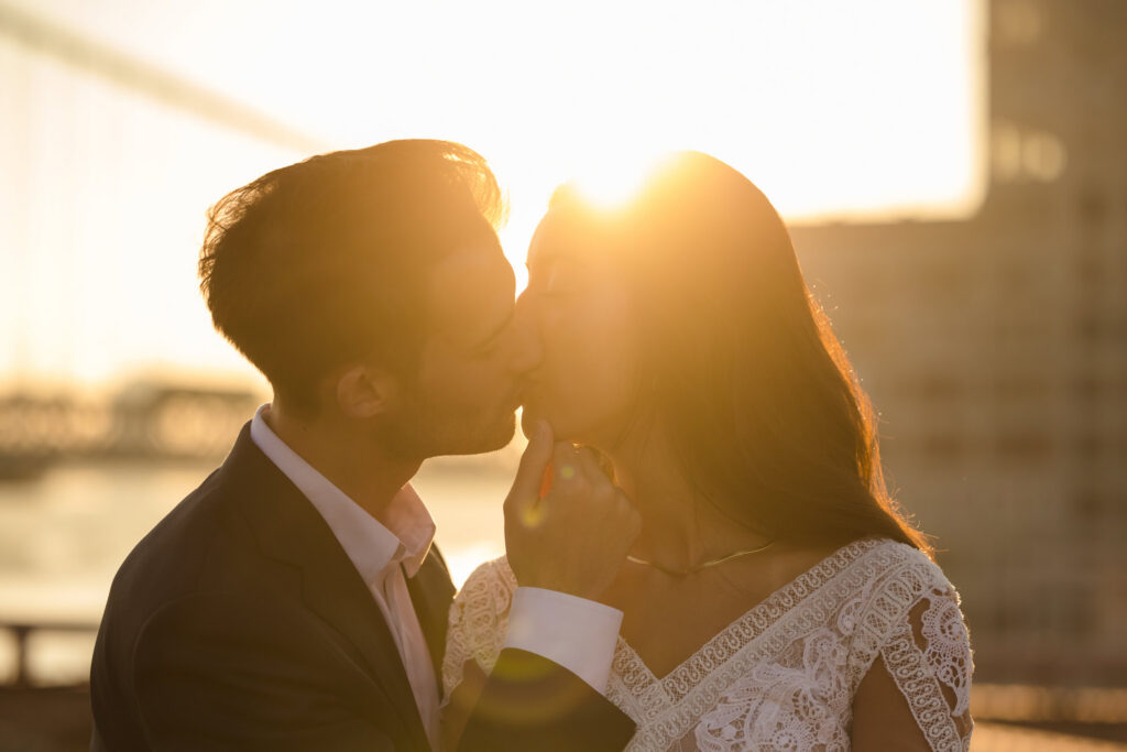 wedding photo of bride and groom kissing in silhouette, backlit by golden July morning sun with lens flare and the Manhattan skyline softly glowing behind them.