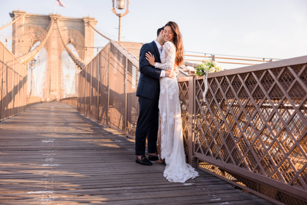 Sunrise Brooklyn Bridge wedding portrait of bride and groom embracing along the bridge railing, bouquet resting nearby, with the iconic stone tower and suspension cables glowing in soft July morning light.