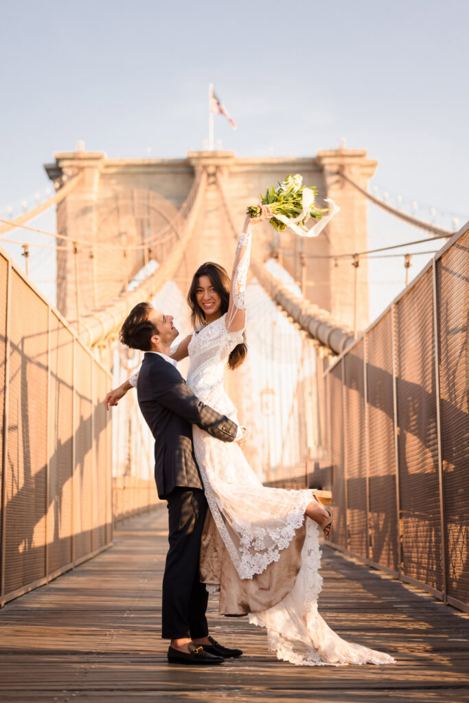 Sunrise Brooklyn Bridge wedding portrait of groom lifting bride as she raises her bouquet overhead, framed symmetrically by the iconic stone arches and suspension cables in warm July morning light.