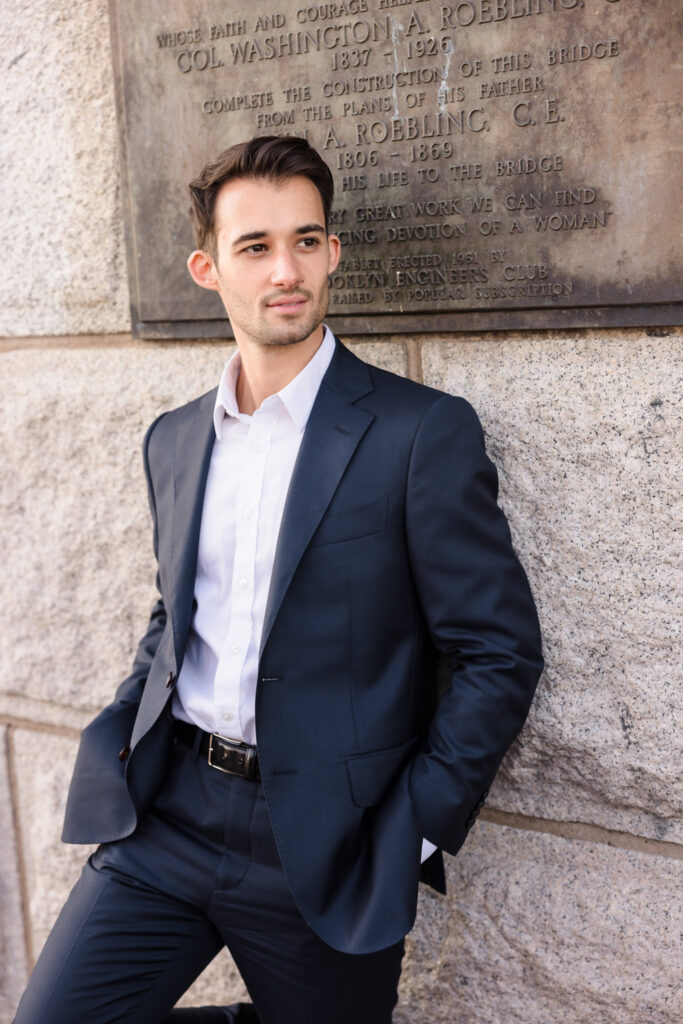Groom portrait at sunrise on the Brooklyn Bridge, standing against the historic Roebling plaque in a navy suit with soft July morning light illuminating the stone architecture.