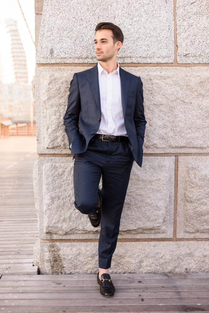 Stylish groom portrait, leaning against the stone pillar in a tailored navy suit with warm July morning light highlighting the historic architecture.