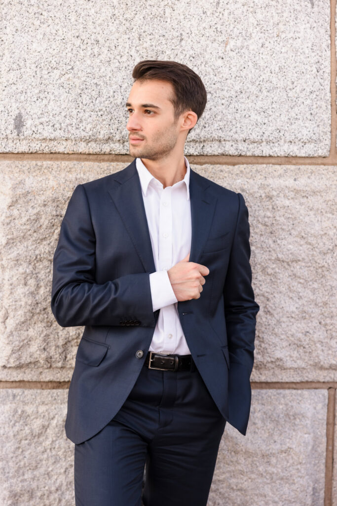 Close-up groom portrait, adjusting his navy suit jacket against the textured stone pillar in soft July morning light.